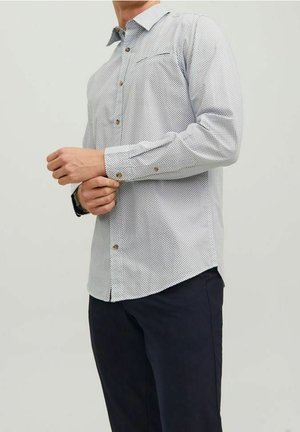 Mann, der die Manschette eines weißen Hemdes mit kleinem schwarzem Rautenmuster zuknöpft, kombiniert mit dunkelblauen Hosen und einer schwarzen Armbanduhr.