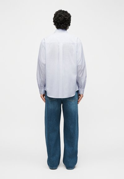 Άτομο με σγουρά μαλλιά που φοράει γαλάζιο ριγέ πουκάμισο με μακριά μανίκια και χαλαρά μπλε τζιν, στέκεται με την πλάτη γυρισμένη σε λευκό φόντο.