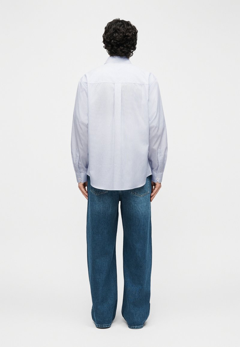 Persoon met krullend haar, gekleed in een lichtblauw gestreept shirt met lange mouwen en losse blauwe spijkerbroek, staand met de rug naar voren op een witte achtergrond.