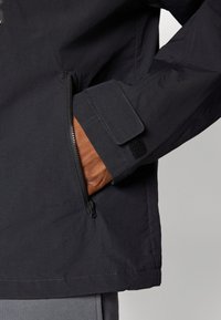 Musta takki, jossa on vetoketjupussi ja tarranauhakä wrist. Ominaisuuksiin kuuluu sileä, kevyt materiaali ja moderni, toiminnallinen muotoilu.