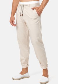 Man met beige drawstring broek opgerold bij de enkels en witte instappers, staande met één hand in zijn zak.