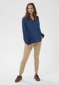 Cămașă albastru închis cu mâneci lungi și decolteu în V, croială lejeră, manșete strânse. Asortată cu pantaloni bej mulați și pantofi maro cu șireturi.