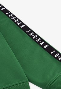 Tejido atlético verde con una superficie texturizada; presenta una cinturilla negra con el logo "JORDAN" en blanco y letras distintivas.