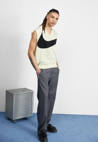 Un pull sans manches vert clair avec un logo Nike noir, associé à un pantalon gris et des chaussures noires. Il présente un design en V et une coupe décontractée.