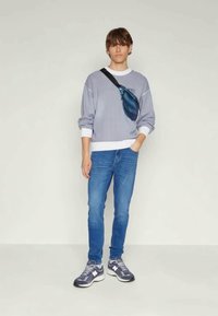 Sivá mikina s bielym lemovaním, modrá crossbody taška, modré džínsy a sivé tenisky. Model stojí vzpriamene pred jednoduchým pozadím.