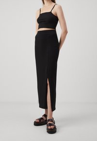 Schwarzes Crop-Top mit dünnen Trägern, kombiniert mit einem langen schwarzen Rock mit einem seitlichen Schlitz; Model trägt klobige schwarze Sandalen.