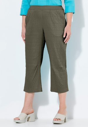 Frau trägt olivgrüne, strukturierte, verkürzte Hose, türkisfarbenes Langarmoberteil und beige Sandalen mit offenem Zehenbereich und klobigem Absatz, steht vor einer weißen Wand.