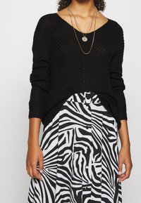 Maglione nero lavorato a maglia con scollatura a V, abbinato a una gonna plissettata bianca e nera a strisce zebrate, con bottoni e vita con cintura.