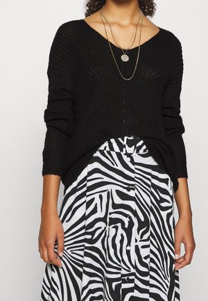 Maglione nero lavorato a maglia con scollatura a V, abbinato a una gonna plissettata bianca e nera a strisce zebrate, con bottoni e vita con cintura.