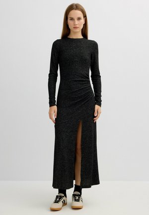 Largo vestido negro en una tela suave y texturada con mangas largas, un corte lateral y detalle de cintura fruncida, combinado con zapatillas de color claro.