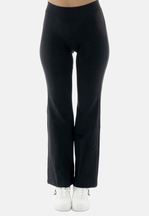 Mujer con pantalones negros acampanados y zapatillas blancas, de pie frente a un fondo claro y liso, recortada a la altura del abdomen y los pies.