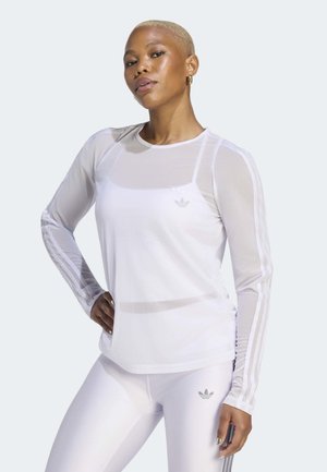 Person med kort blondt hår iført en hvid langærmet mesh-top og hvide leggings med Adidas-logoer poserer med den ene hånd på hoften.