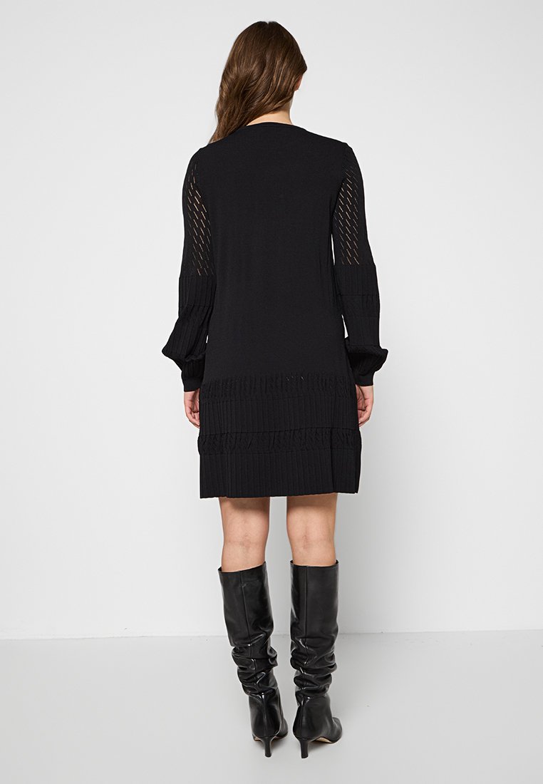 Femme portant une robe en tricot noire à manches longues avec des panneaux de manches transparentes et des bottes noires montantes et décontractées, se tenant devant un fond uni.