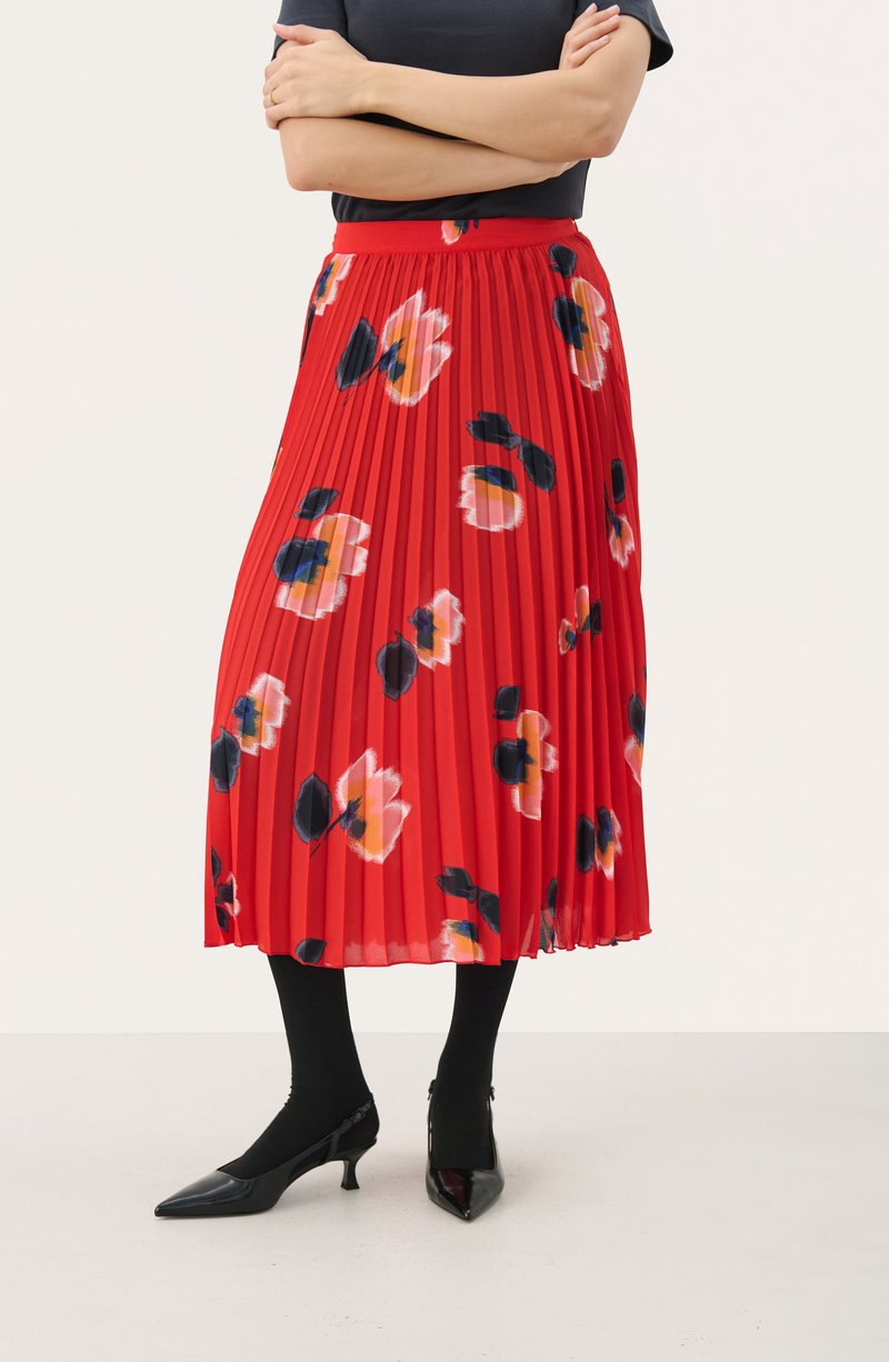 Rød plisseret nederdel med et blomstermønster i pink, sort og orange. Parret med sorte strømpebukser og spidse hæle. Glat, let stof.