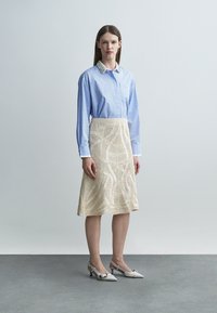 Kvinne som står iført blåstripet skjorte med pyntet krage, kremfarget teksturert knelang skjørt og hvite lave hæler.