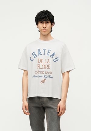 Jeune homme aux cheveux noirs portant un t-shirt blanc oversize avec un texte bleu et orange et un jean gris, debout devant un fond blanc uni.