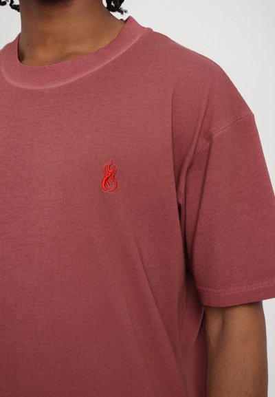 Camiseta de manga corta y cuello redondo en un color burdeos apagado. Presenta un pequeño bordado de llama roja en el pecho. Textura suave, ajuste relajado.