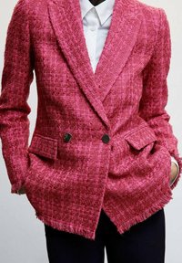 Vaaleanpunainen tweed-bleiseri, jossa on tekstuuri. Kaksoisnapitus, kaksi etutaskua ja purjehuppumaiset helmat. Päällä valkoinen paita.