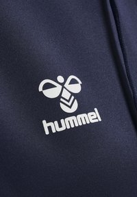 Białe logo Hummel ze stylizowaną pszczołą i napisem na granatowym materiale, prawdopodobnie sportowa odzież.