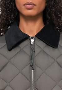 Gevoerd grijs jack met een zwarte corduroy kraag, voorzien van een ritssluiting en trekkoorddetails bij de nek. Gladde textuur.