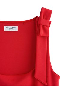 Červený tkaninový top s texturovaným povrchem. Má mašličku na rameni. Štítek zní "Holly & Whyte by Lindex," vyrobeno v Turecku, velikost EUR S UK S.