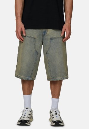 Osoba ubrana w wyblakłe dżinsowe szorty do kolan, białe skarpetki sportowe i białe buty sportowe z czarnymi elementami, stojąca z rękami swobodnie opuszczonymi wzdłuż ciała.