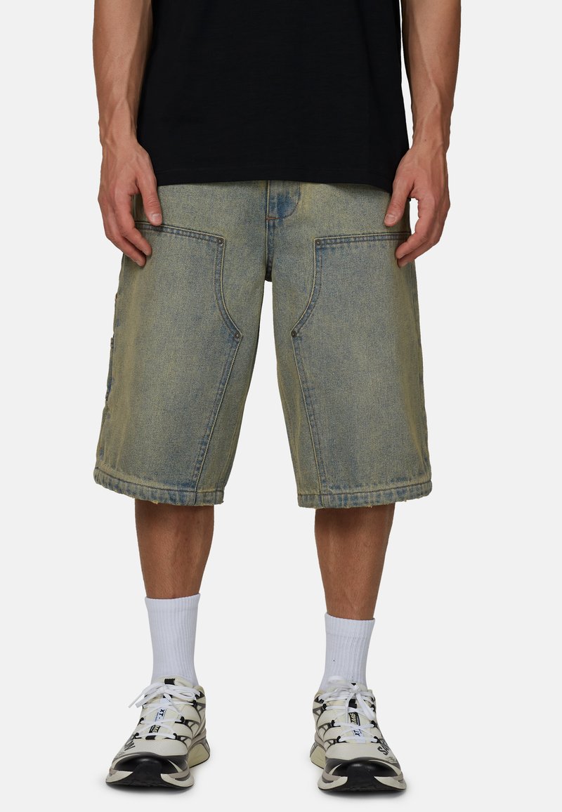 Osoba ubrana w wyblakłe dżinsowe szorty do kolan, białe skarpetki sportowe i białe buty sportowe z czarnymi elementami, stojąca z rękami swobodnie opuszczonymi wzdłuż ciała.