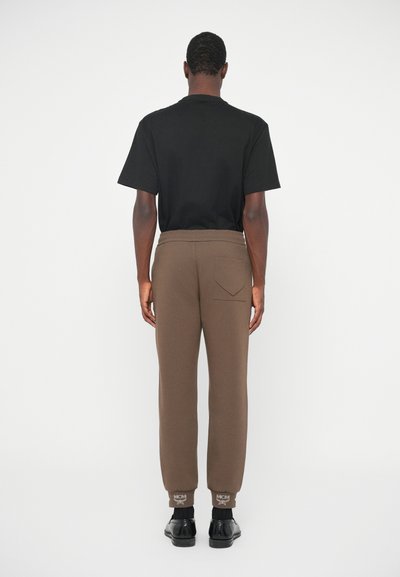 Man staat met zijn rug naar de camera, gekleed in een zwart T-shirt, bruine joggingsbroek met achterzak, zwarte sokken en zwarte dresschoenen, op een witte achtergrond.