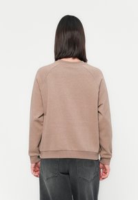 Sweatshirt marron avec un col rond, des manches raglan et un ourlet côtelé. Porté avec un jean foncé légèrement usé doté d'un logo sur la poche arrière.