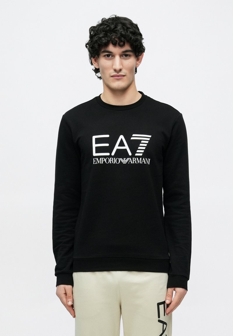 Fiatal férfi göndör hajjal, fekete EA7 Emporio Armani pulóvert és bézs nadrágot visel, melyen az EA7 logó található a lábszárnál, fehér háttér előtt áll.