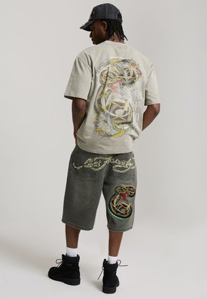 Mężczyzna w beżowej koszulce z dużą kolorową grafiką węża oraz szarych szortach z haftowanym wężem i stylizowanym tekstem na plecach.