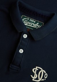 Dunkelblaues Poloshirt mit strukturiertem Stoff, drei weißen Knöpfen am Kragen und einem in Creme gehaltenen Sticklogo auf der linken Seite.