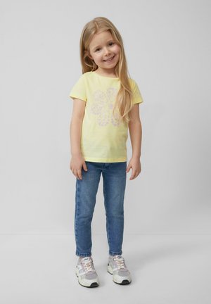 Gelbes Baumwoll-T-Shirt mit kurzen Ärmeln und pinkem Blumenmuster, kombiniert mit blauen Jeans und mehrfarbigen Turnschuhen. Heller Hintergrund.