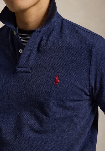 Bărbat care poartă un tricou polo bleumarin cu guler ridicat și un logo roșu brodat pe piept, arătând gâtul și partea superioară a trunchiului.