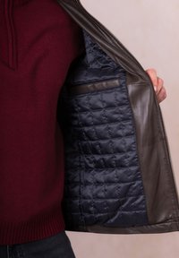 Dark brown leather jacket with a quilted navy interior, featuring a zip closure and a pocket inside. Worn over a maroon sweater.