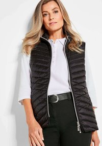 Schwarze gesteppte Weste mit hohem Kragen und Frontreißverschluss, über einem weißen Polo-Shirt getragen. Ein schwarzer Gürtel sorgt für einen Kontrast zum Outfit.
