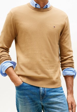 Homme portant un pull beige col rond sur une chemise bleue à rayures et boutons avec manches retroussées, associé à un jean bleu, main dans la poche.