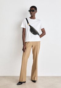 Biała bawełniana koszulka, beżowe dopasowane spodnie i czarna torba na ramię z złotymi akcentami. Model nosi czarne okulary przeciwsłoneczne i czarne buty na spiczastym nosku.