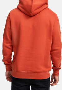 Orange bomullshoodie med huva, långa ärmar och ribbat avslut. Slät textur, inga synliga mönster eller hårdvara, visas från bakvyn.