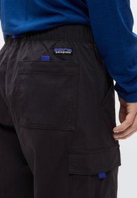 Κοντινό πλάνο ενός ατόμου που φοράει μαύρο παντελόνι Patagonia με ελαστική ζώνη στη μέση και πολλαπλές τσέπες, συνδυασμένο με μπλε πουκάμισο μακρυμάνικο.