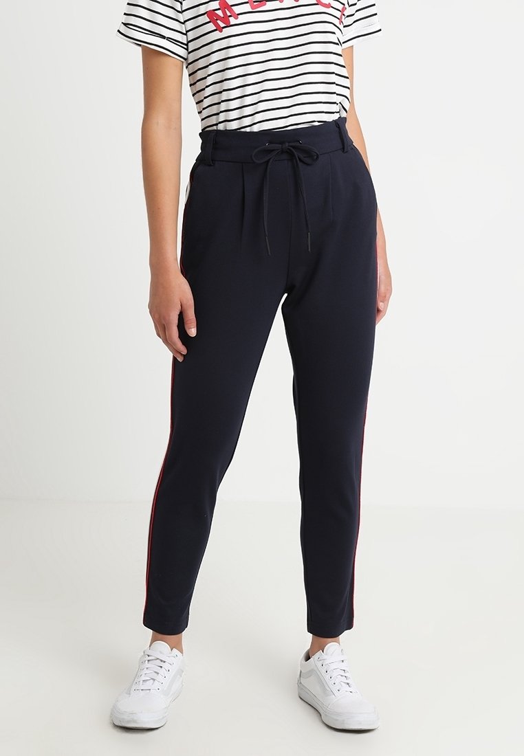Personne debout portant un t-shirt à rayures, un pantalon bleu marine à cordon avec des bandes rouges sur les côtés, et des baskets blanches dans une pièce claire et simple.