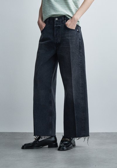 Persoon draagt zwarte wijde jeans met onafgewerkte zomen, gecombineerd met zwarte leren loafers en een groen gestreept overhemd, staand op een grijze vloer.