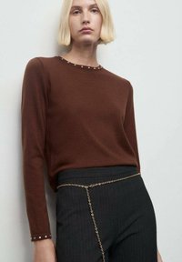 Una donna con i capelli biondi corti indossa una maglietta marrone a maniche lunghe con piccoli rivetti sui polsini e sul collo, pantaloni neri e una cintura a catena d'oro.