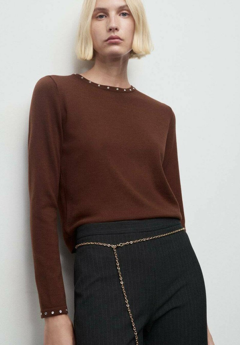 Una donna con i capelli biondi corti indossa una maglietta marrone a maniche lunghe con piccoli rivetti sui polsini e sul collo, pantaloni neri e una cintura a catena d'oro.