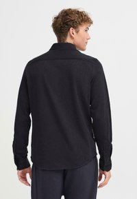 Camicia nera a maniche lunghe con colletto, caratterizzata da una texture liscia e polsini abbottonati. Il retro presenta un orlo curvo e cuciture sulle spalle.