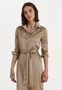 Beige overhemdjurk met een kraag, knoopsluiting aan de voorkant en opgerolde mouwen. Heeft een gestrikte taille en een herhalend geometrisch patroon in een donkerdere tint.