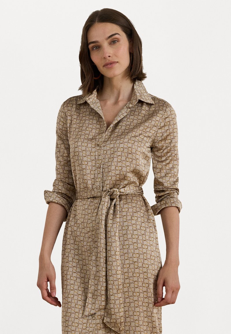 Beige overhemdjurk met een kraag, knoopsluiting aan de voorkant en opgerolde mouwen. Heeft een gestrikte taille en een herhalend geometrisch patroon in een donkerdere tint.
