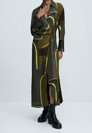 Osoba ubrana w długą, wzorzystą sukienkę kopertową w odcieniach oliwkowego, czarnego i żółtego, z czarnymi masywnymi botkami za kostkę, stojąca na jednolitym tle.