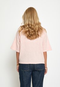 Gaišs rozā, īsām piedurknēm džemperis, izgatavots no mīksta, pūkaina materiāla, ar brīvu piegriezumu un taisnu apakšmalu. Kombinēts ar tumši ziliem džinsiem.