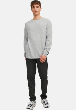 Ung mand, stående, iført en lysegrå langærmet sweater, sorte bukser og hvide sneakers med sorte detaljer, på en ensfarvet hvid baggrund.